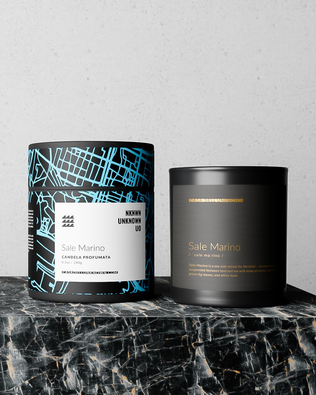 Two candles from the brand Casa dell' Unknown named 'Sale Marino', displayed on a black marble surface against a light gray textured wall.